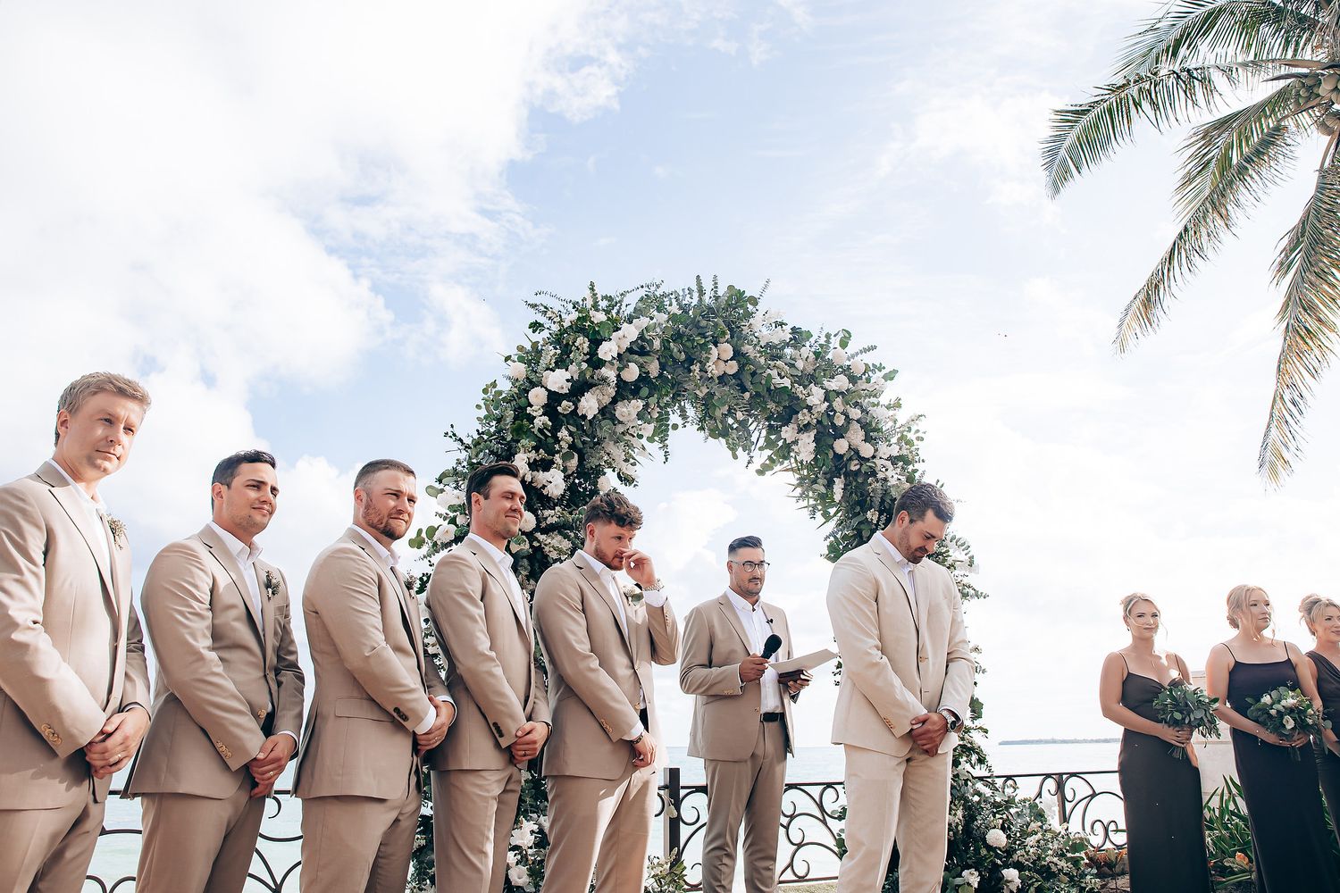 Groomsmen in tan suits line up outdoors with palm trees in the background.