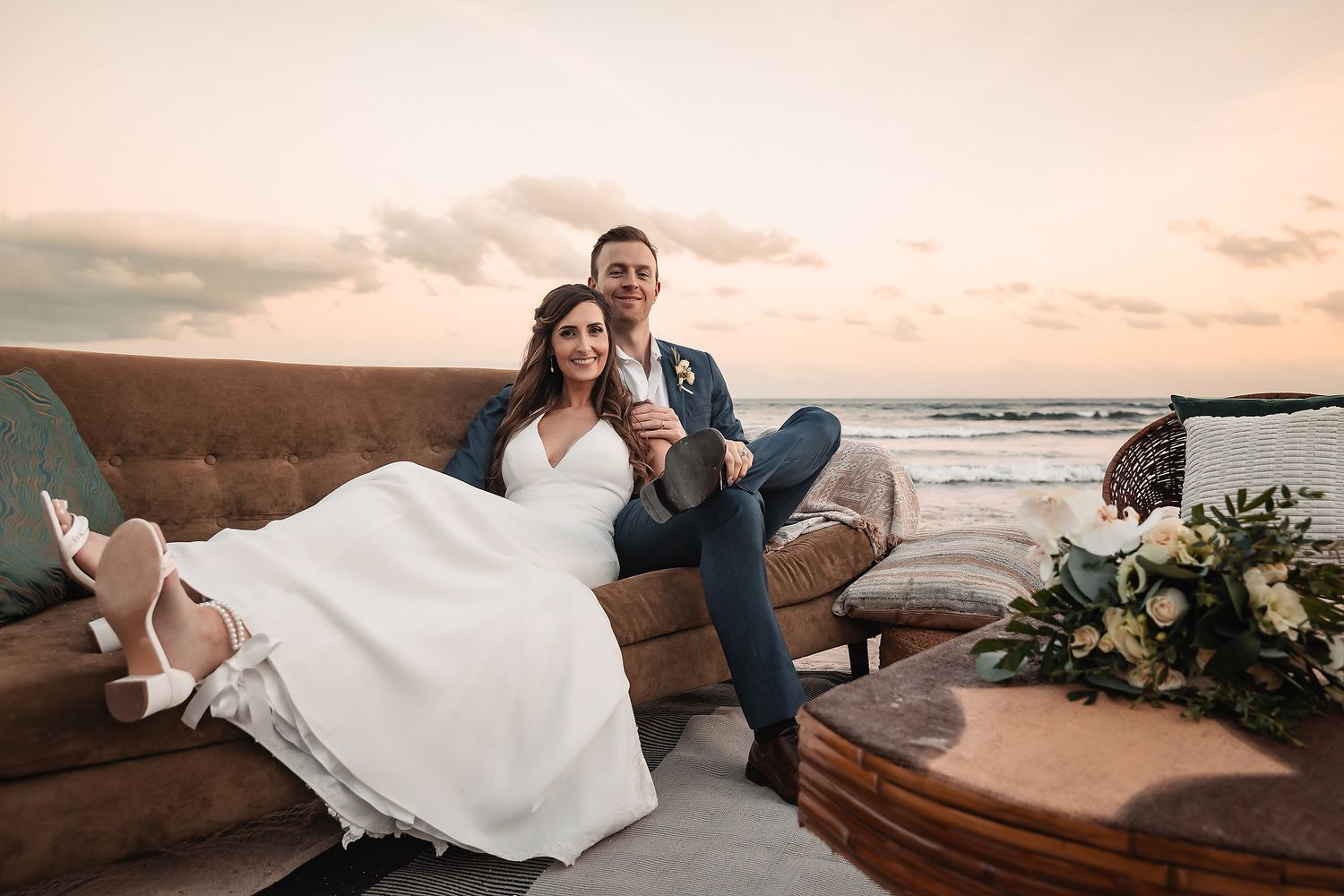 Couple relaxes on a brown leather couch at sunset during a beach wedding photoshoot.