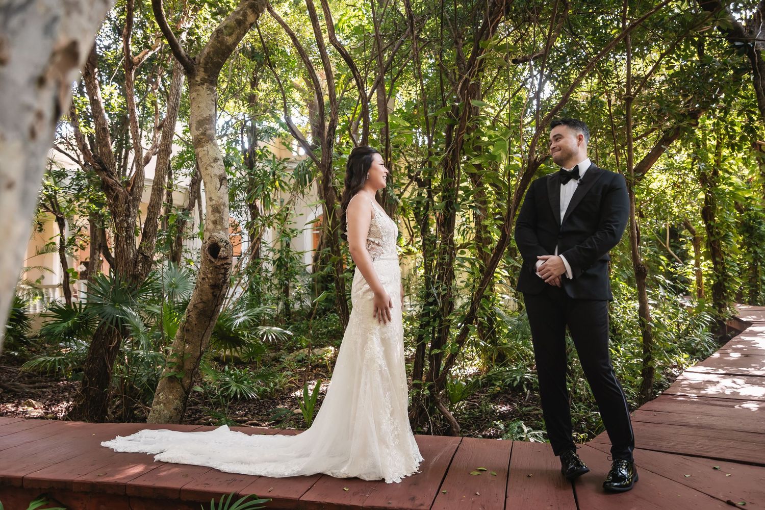 A wedding couple shares a tender moment on a wooden walkway surrounded by lush tropical foliage.