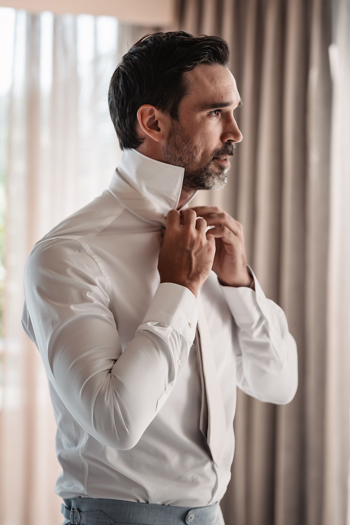 Person in crisp white dress shirt adjusting collar near window with curtains.