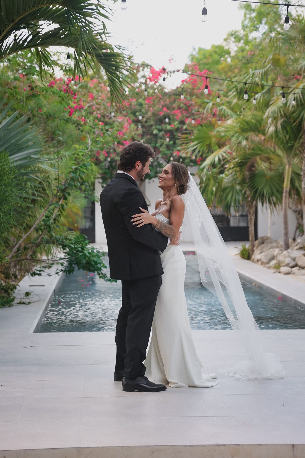 Bride and groom share a romantic moment by a pool, surrounded by tropical palms and pink bougainvillea at an outdoor wedding.