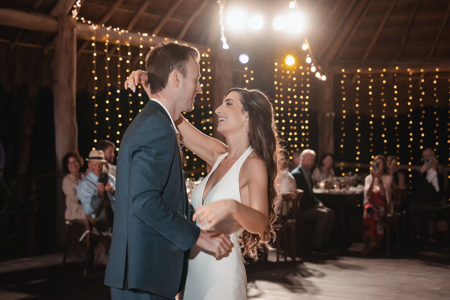 Bride and groom share their first dance under warm string lights in a rustic barn wedding reception.