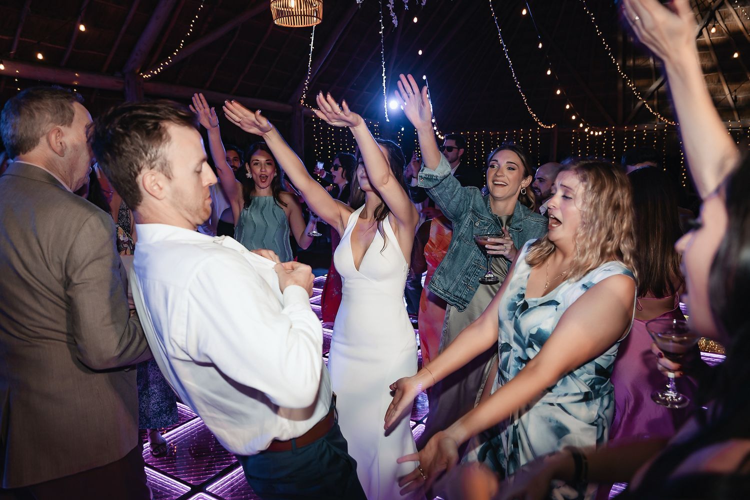 Wedding guests dancing joyfully at a lively reception party with string lights and colorful stage lighting.