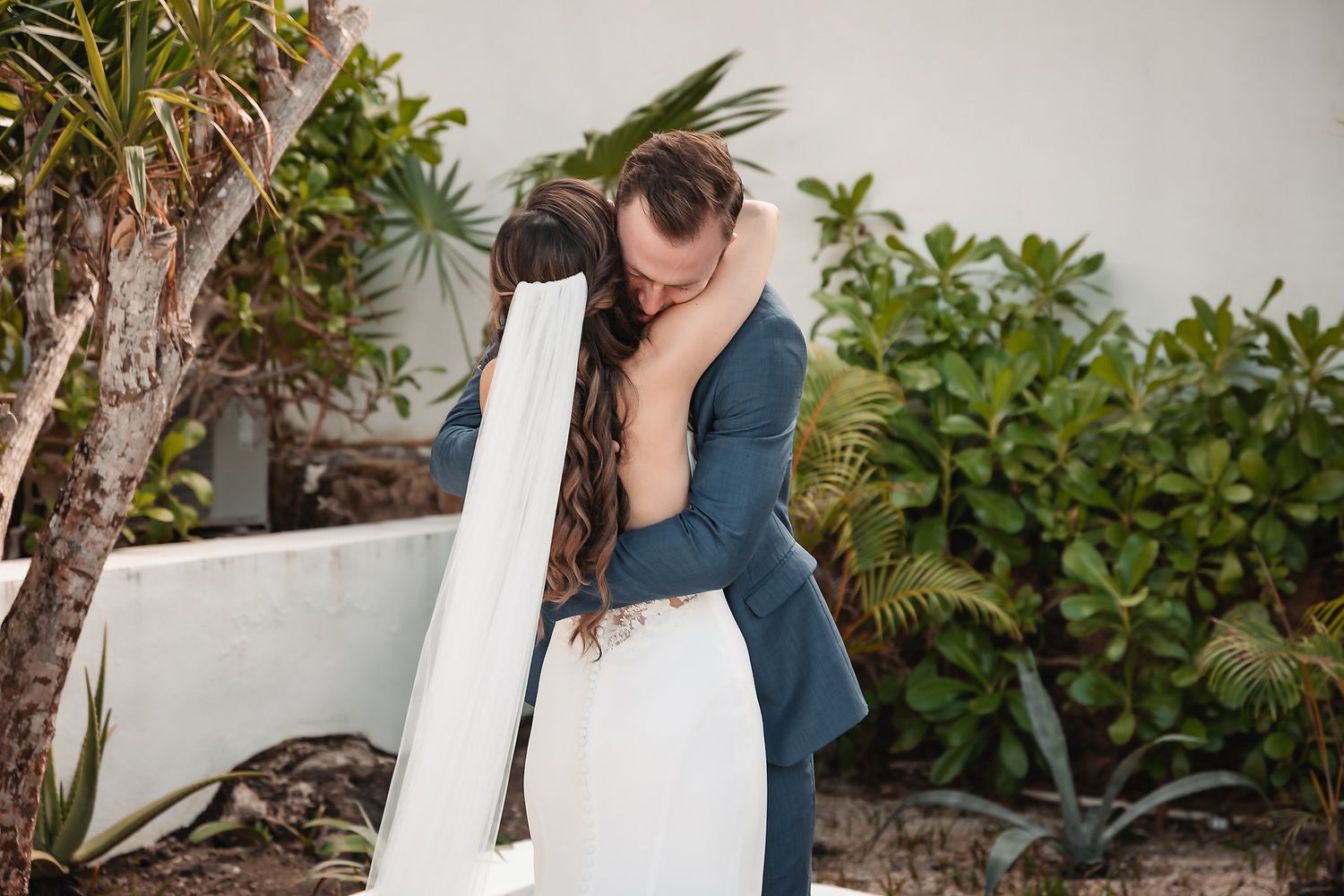 A bride and groom share a tender embrace outdoors, surrounded by lush tropical greenery on their wedding day.