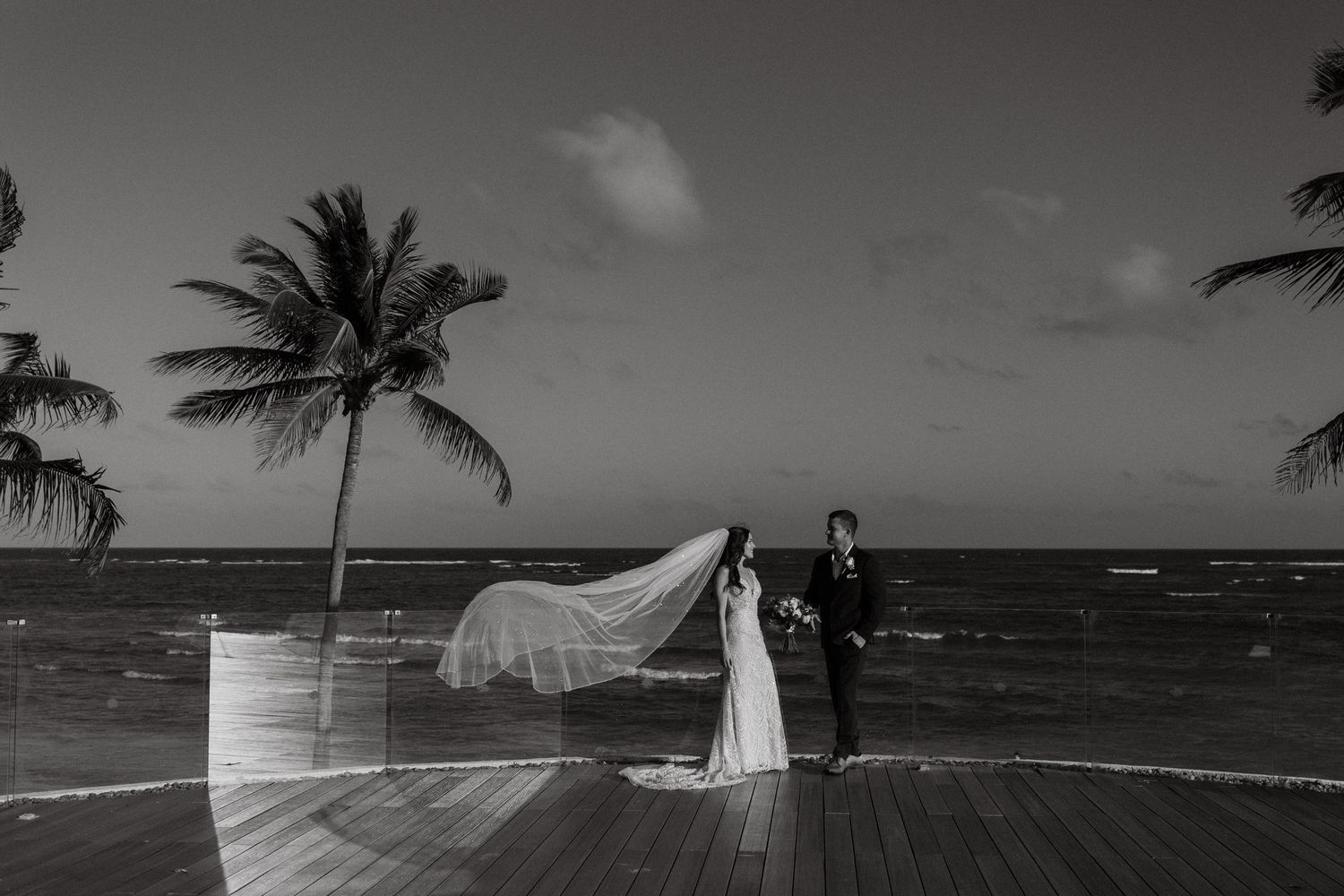 Black and white wedding photo of bride and groom on oceanfront deck with palm trees and flowing veil.