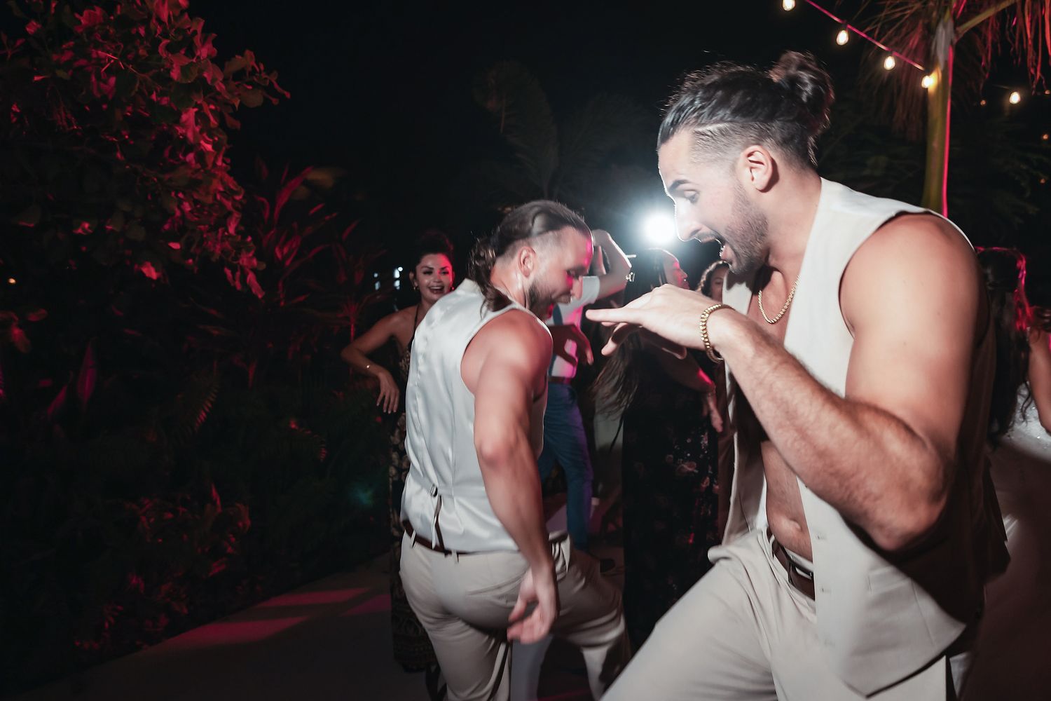 Two men dancing energetically at an outdoor nighttime party, wearing white tank tops and khaki pants.