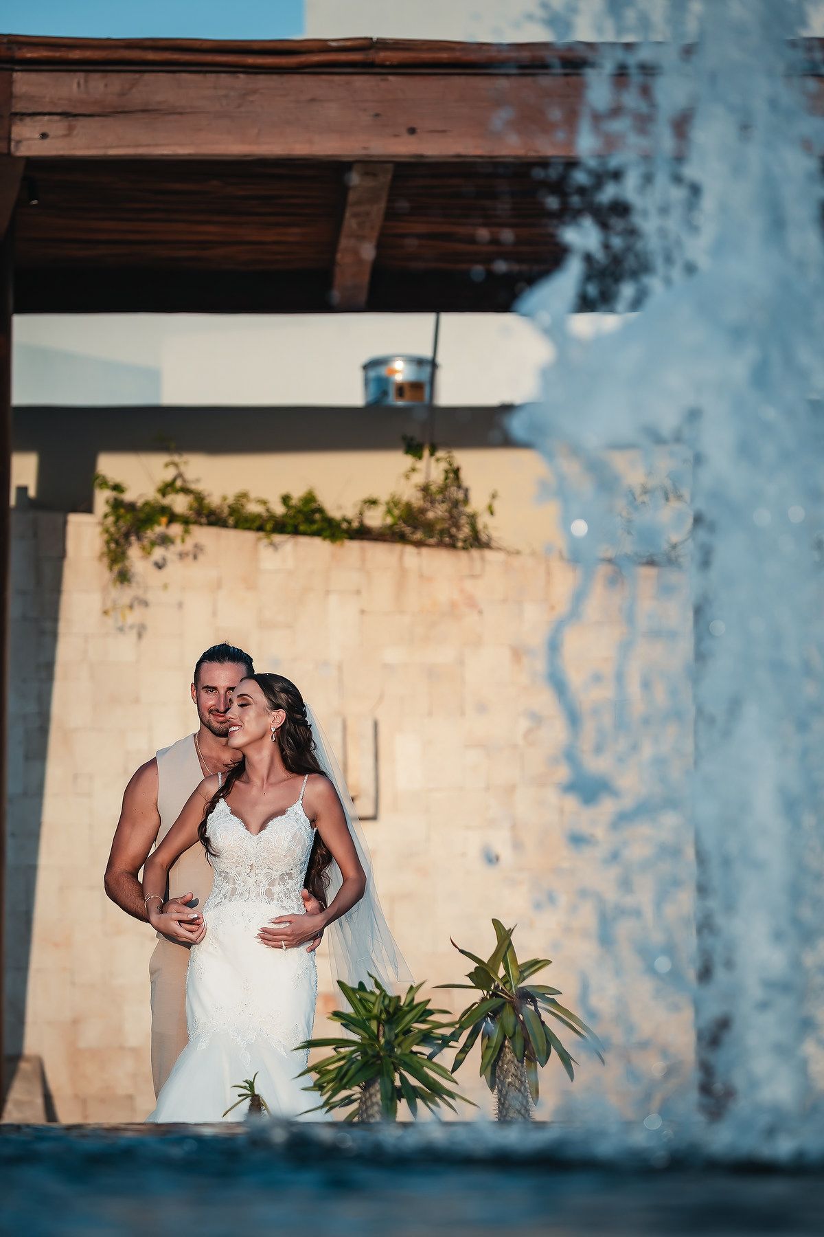 Bride and groom embracing near a fountain at golden hour, with lush greenery and stone wall backdrop.
