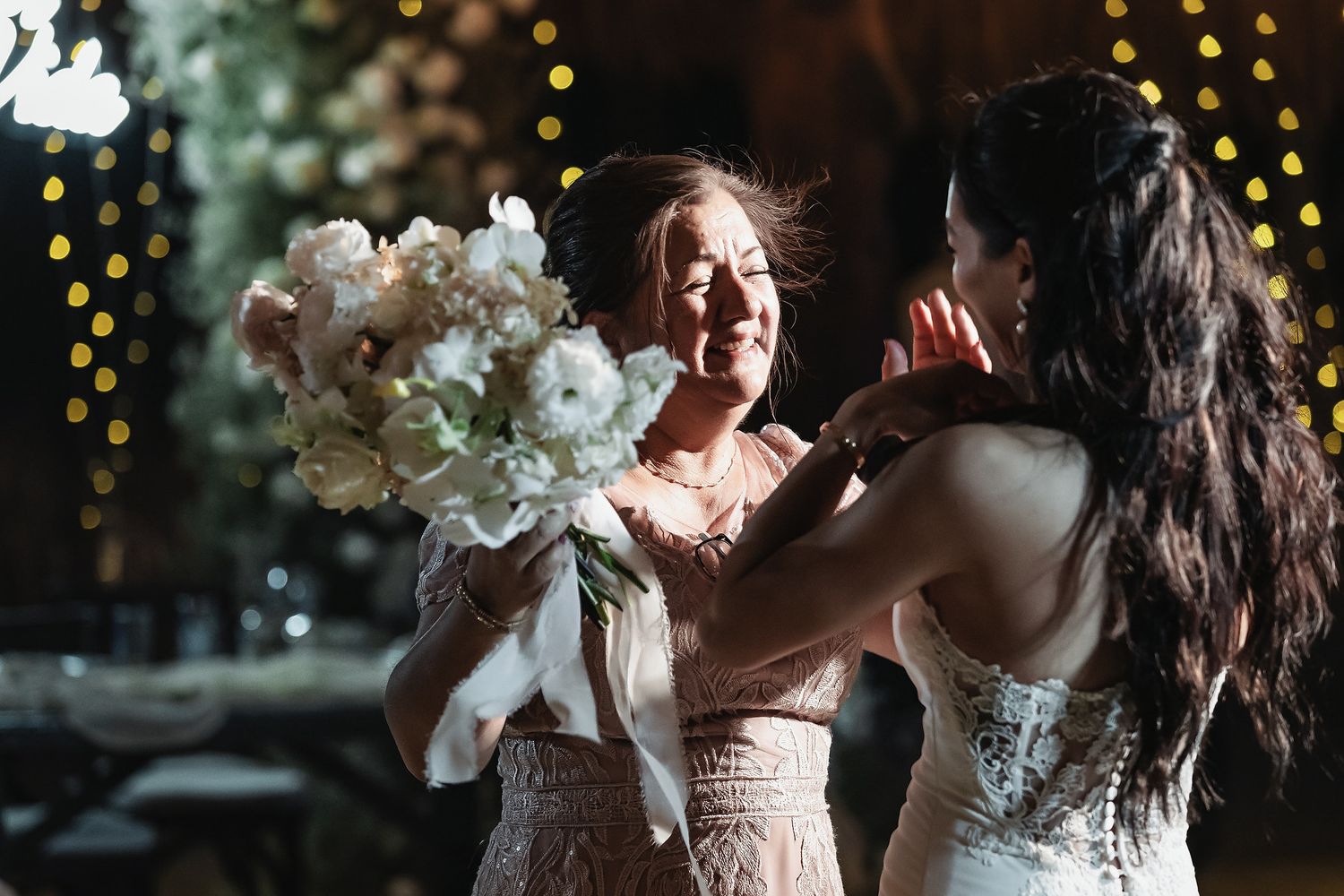 Bride and bridesmaid share emotional moment, holding white hydrangea bouquet with bokeh lights in background.