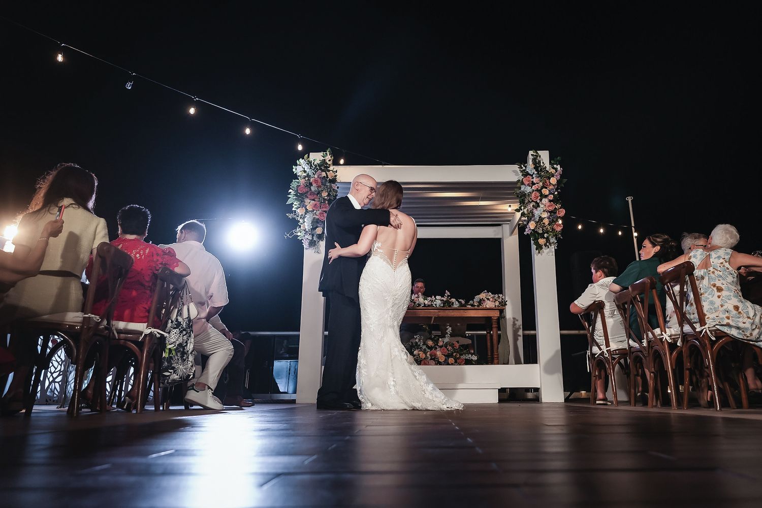 Bride and groom share first dance under floral arch at romantic outdoor wedding reception with string lights.