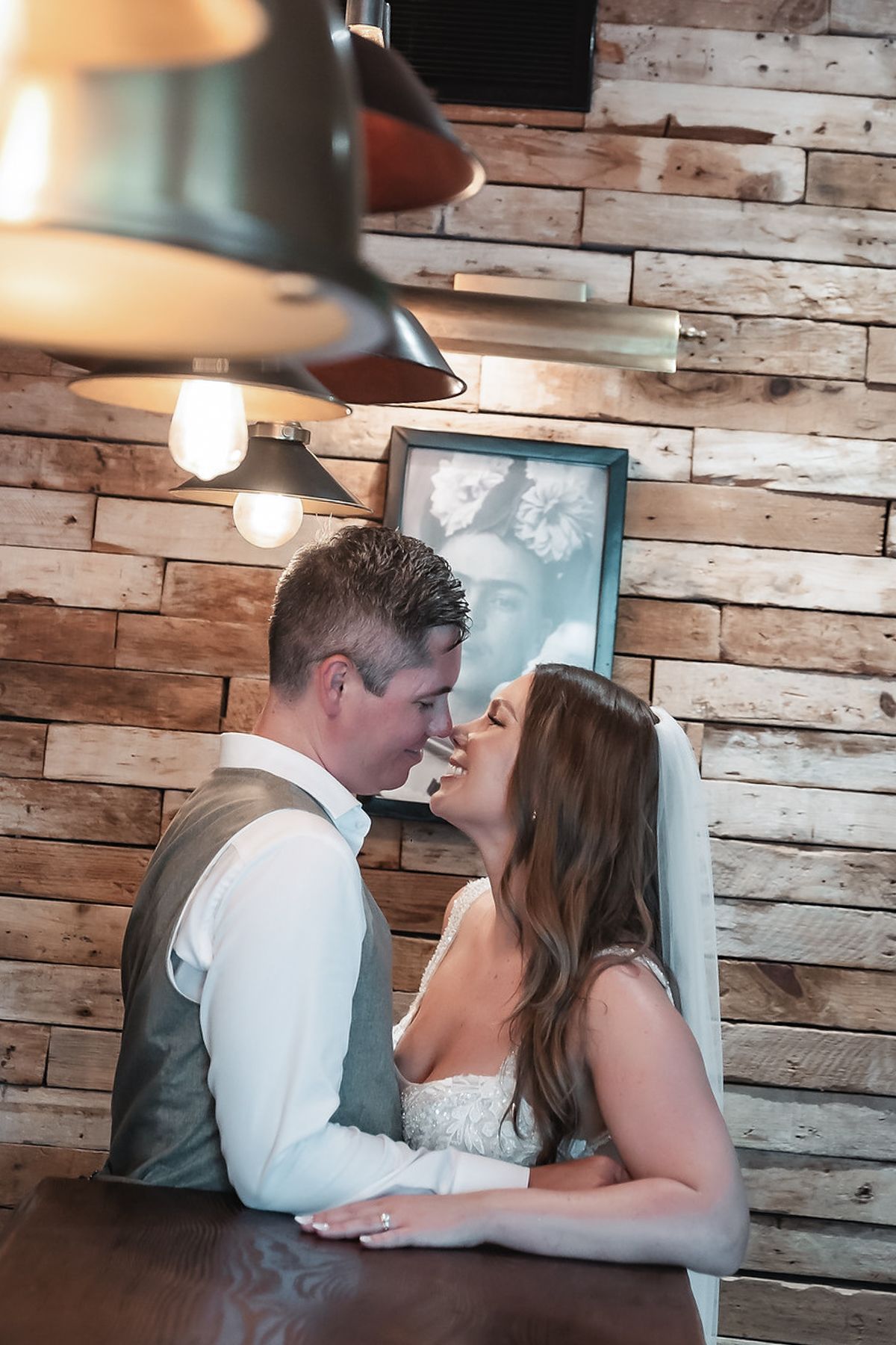 Bride and groom share a romantic moment at a rustic wooden wall venue, soft Edison bulb lighting overhead.