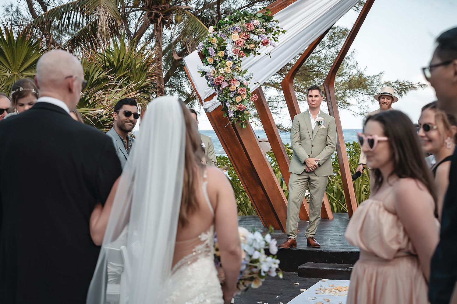 Outdoor wedding ceremony with bride in white veil facing groom at floral arch, guests and bridesmaids in pink dresses surrounding them.
