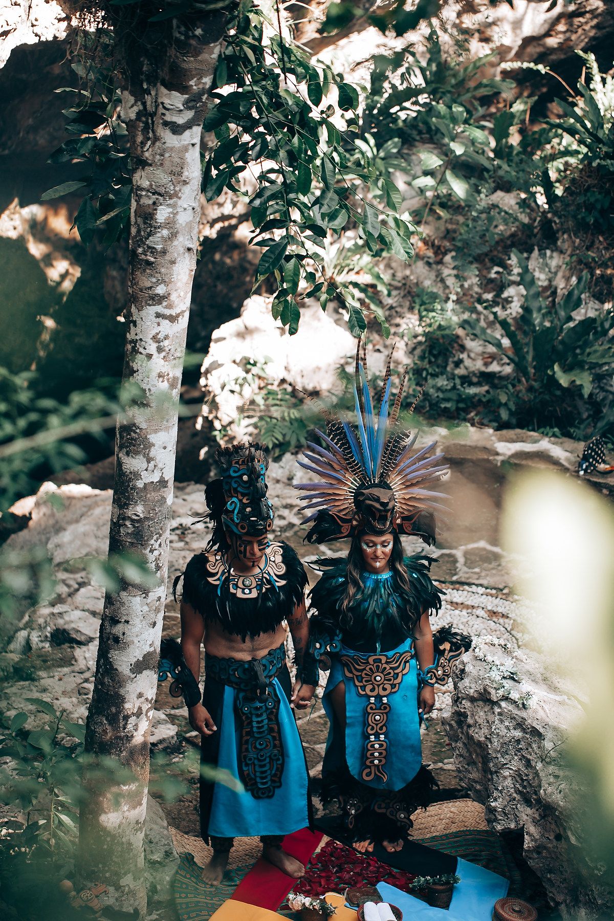 Two people in traditional Mayan ceremonial costumes with turquoise and feather headdresses stand in a jungle setting.