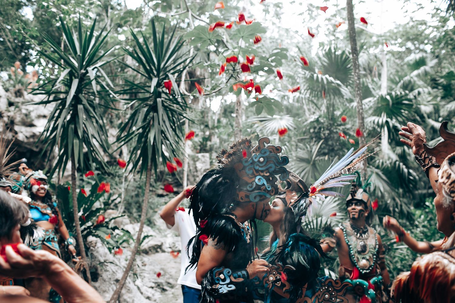 Couple in Mayan ceremonial costumes kissing amid falling red and white flower petals in a jungle.
