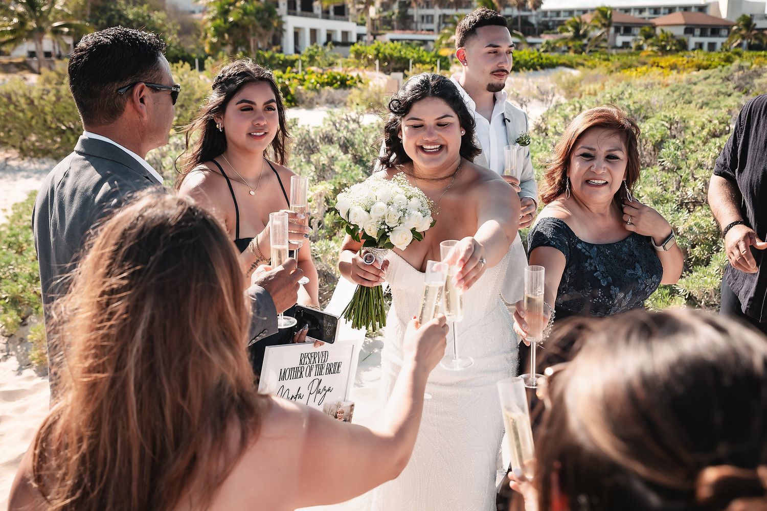 Bride in white gown holds bouquet, toasting with guests outdoors at a sunny beach wedding celebration.