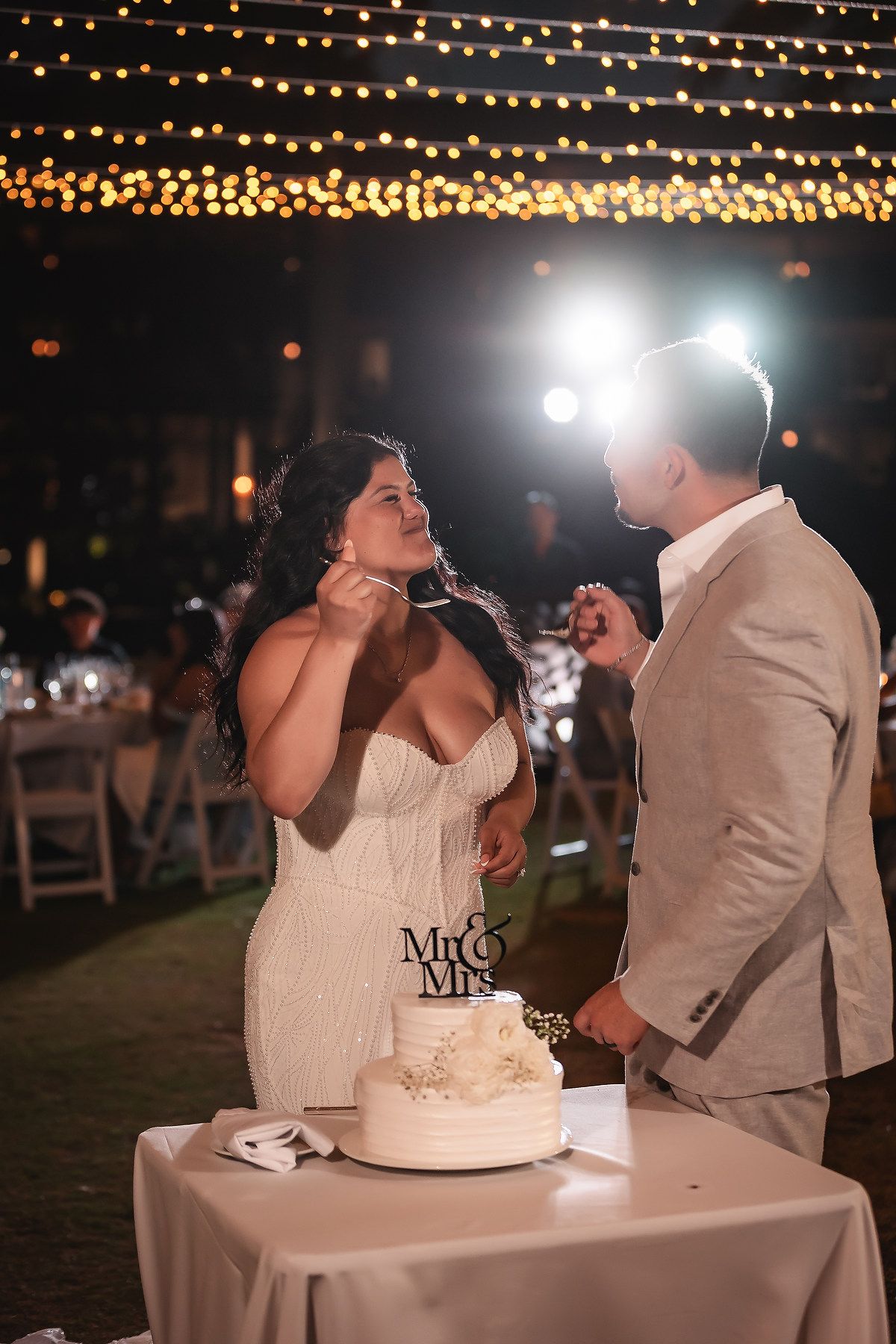 Bride and groom feed each other wedding cake beside a white tiered cake with Mr. & Mrs. topper under string lights.