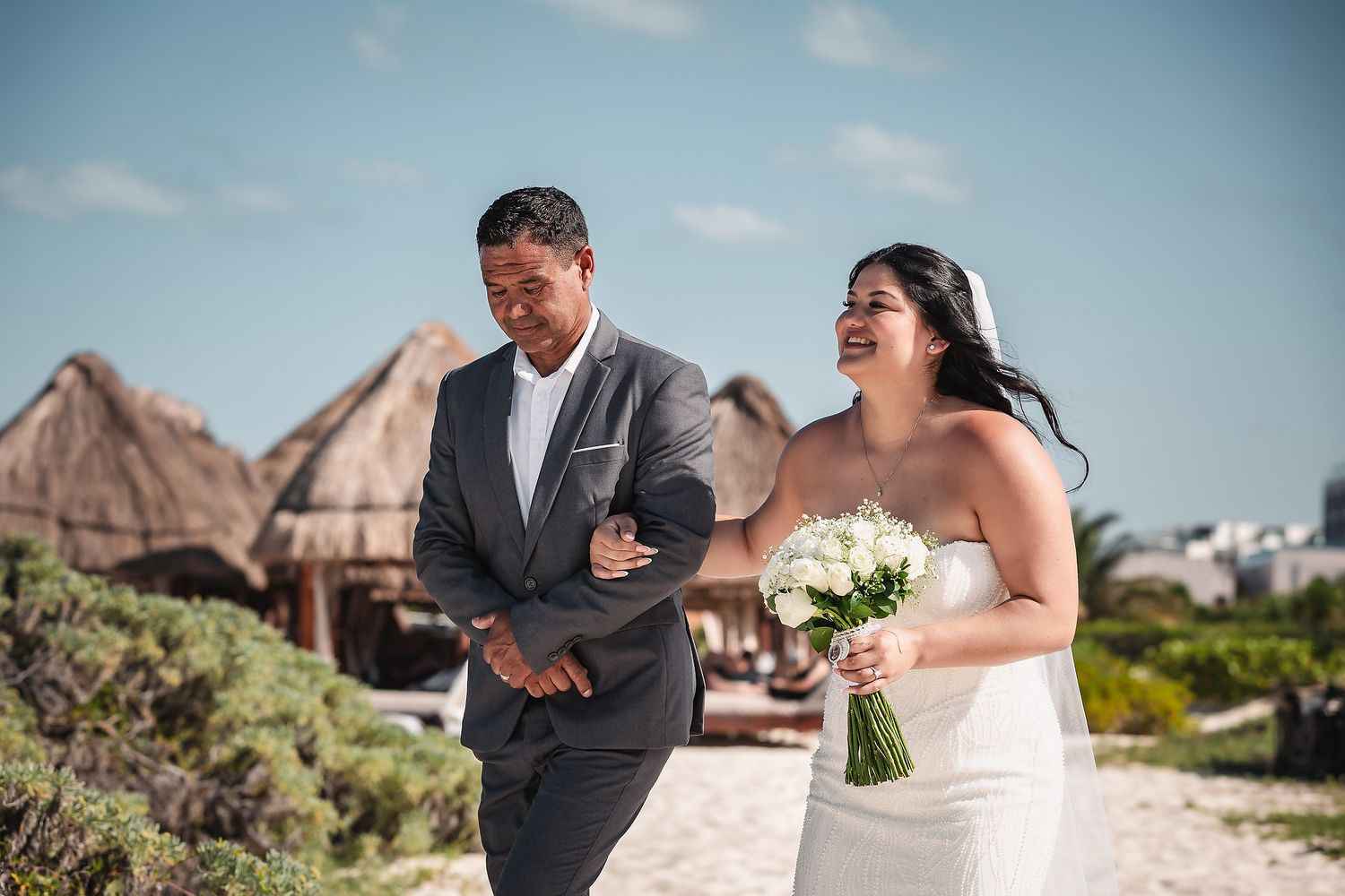 Bride and groom walking together on a sunny beach, bride holding white bouquet, thatched huts in background.