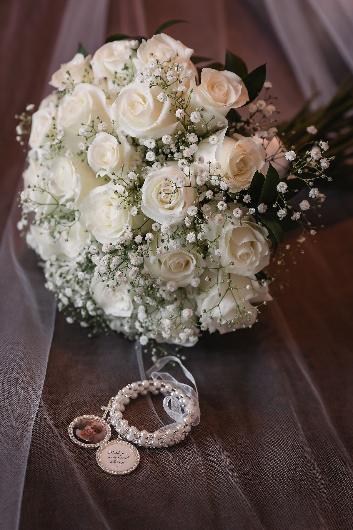 Elegant bridal bouquet of white roses and baby's breath with pearl bracelet and locket on sheer fabric.