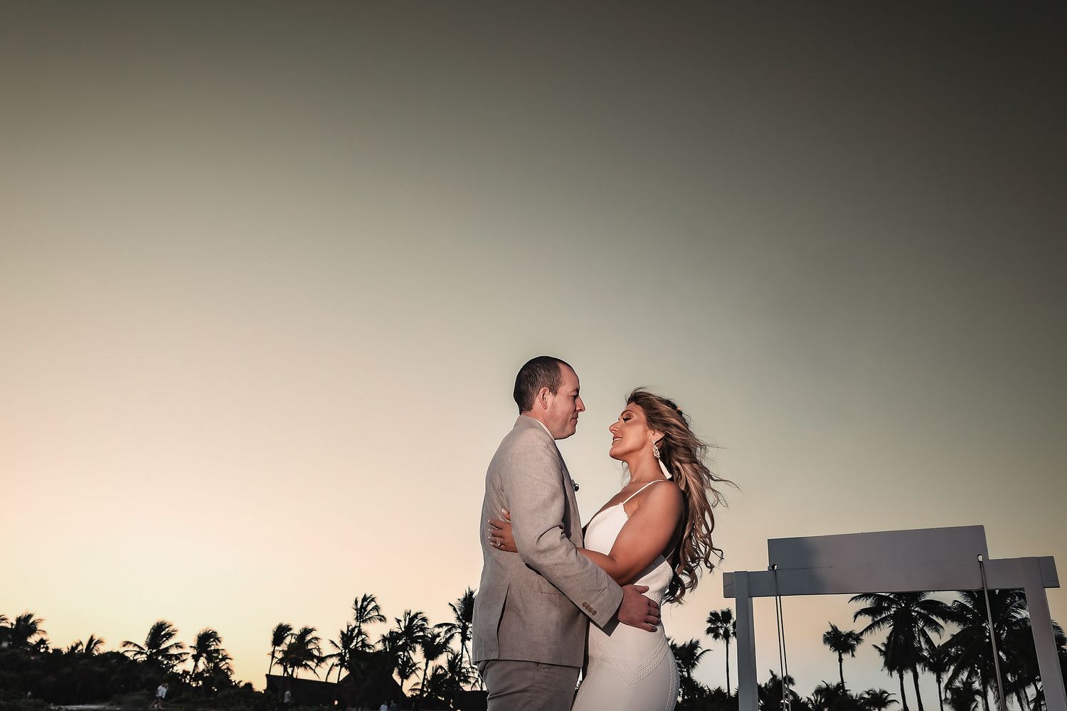 Couple embraces at sunset with palm trees backdrop at tropical beach wedding venue.