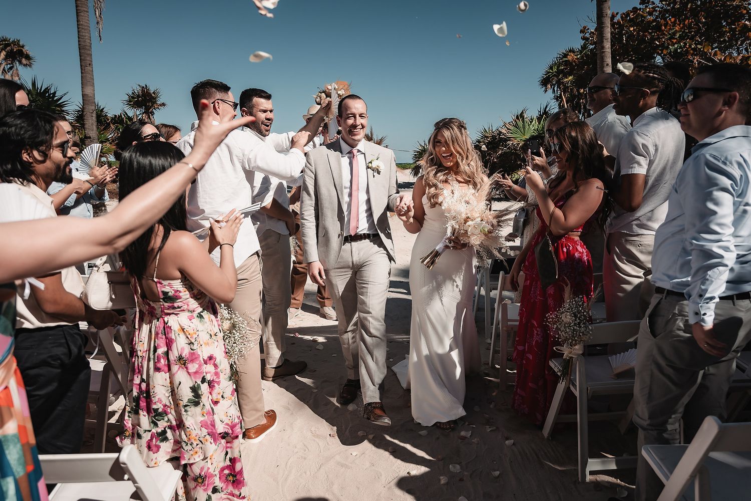 Joyful newlyweds walk down beach aisle as guests toss flower petals during sunny outdoor wedding ceremony.