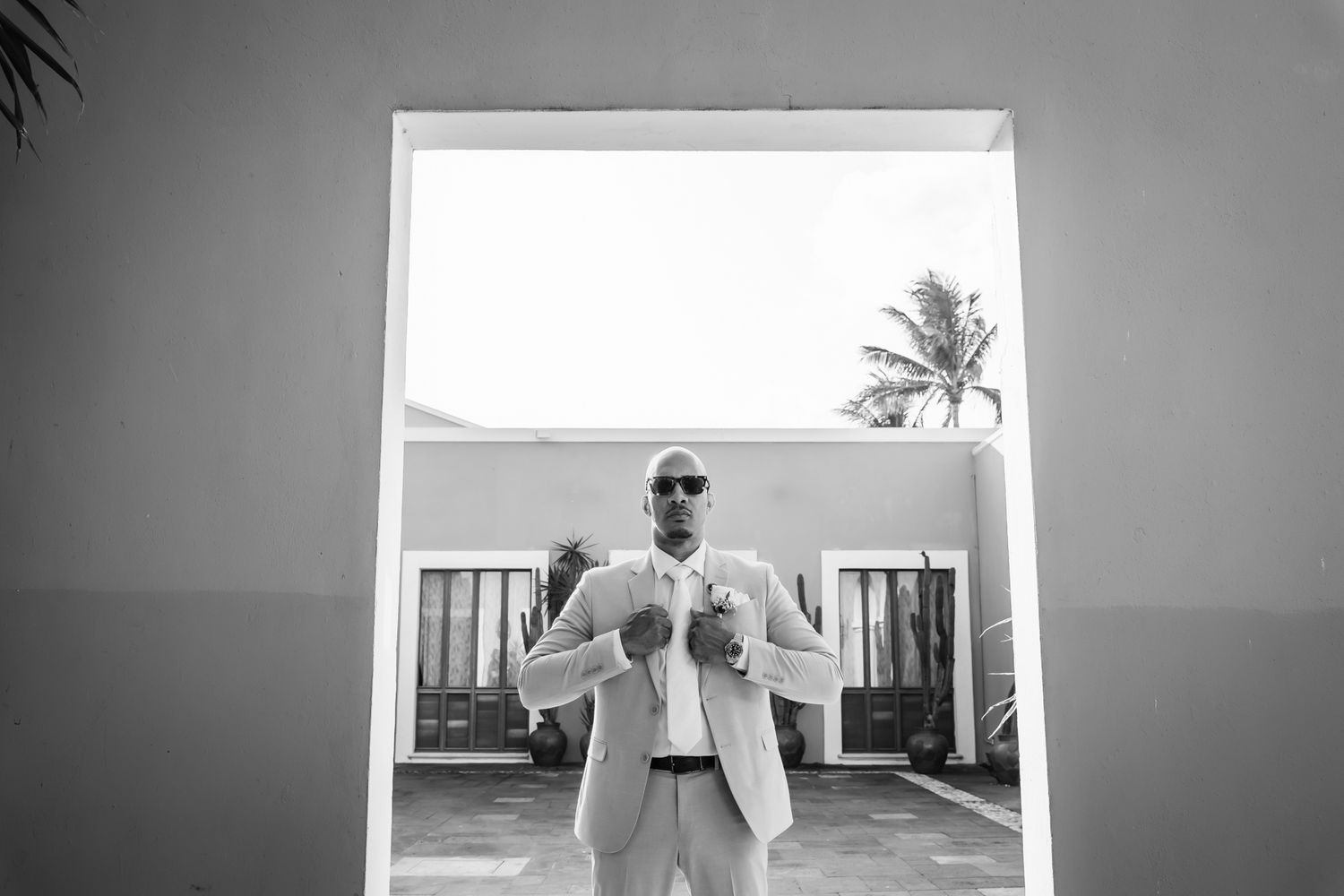 Black and white photo of a bald man in a light suit and sunglasses standing in a doorway with palm trees behind him.
