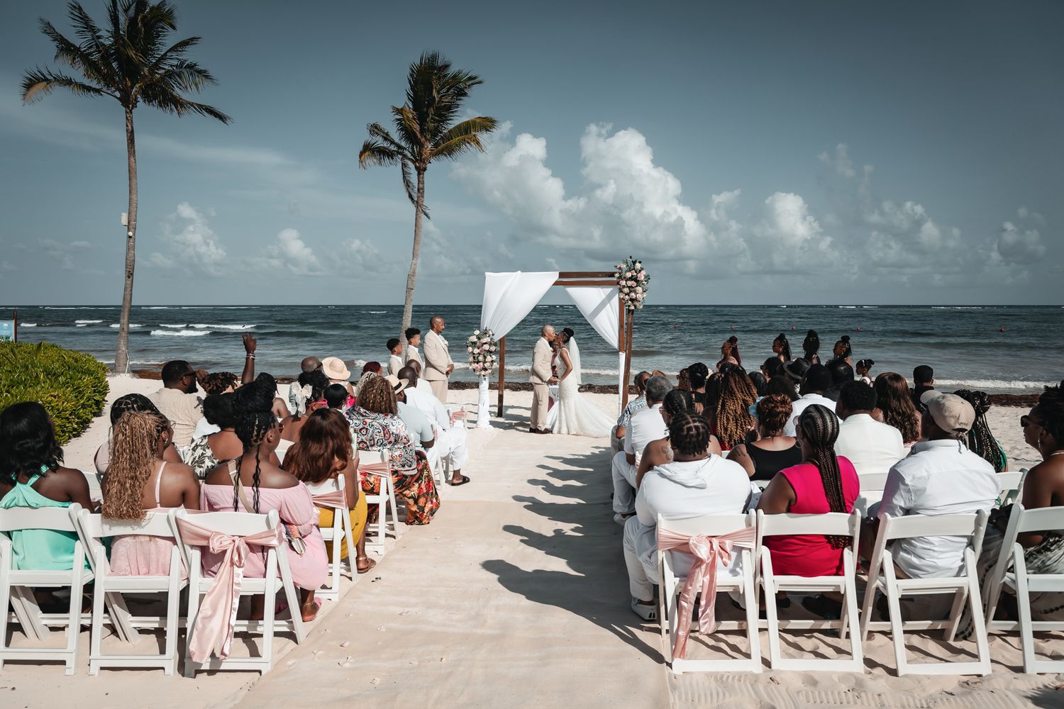 Outdoor beach wedding ceremony with guests seated in white chairs facing an altar adorned with a wooden arch by the ocean.