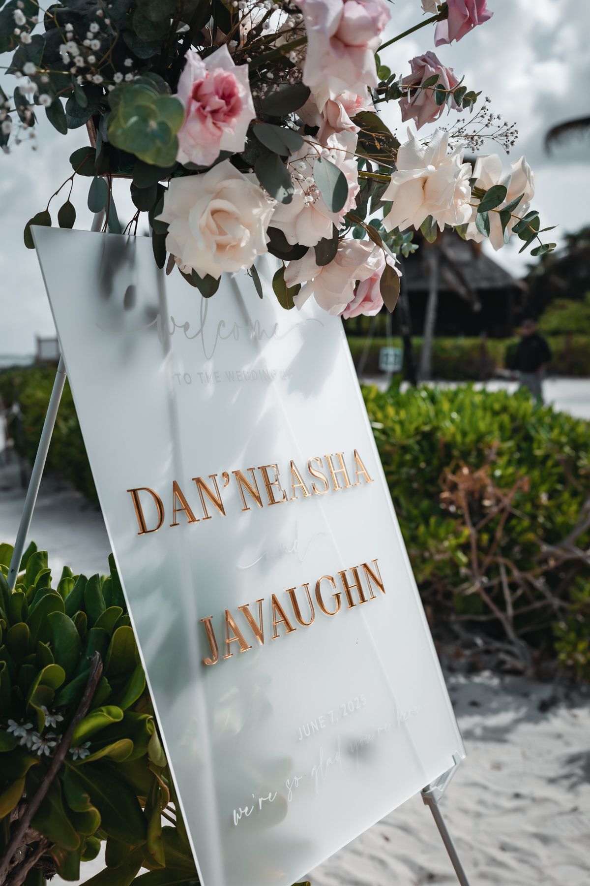 Elegant frosted acrylic wedding welcome sign for Dan'Neasha and Javaughn, adorned with pink roses on a sandy beach.