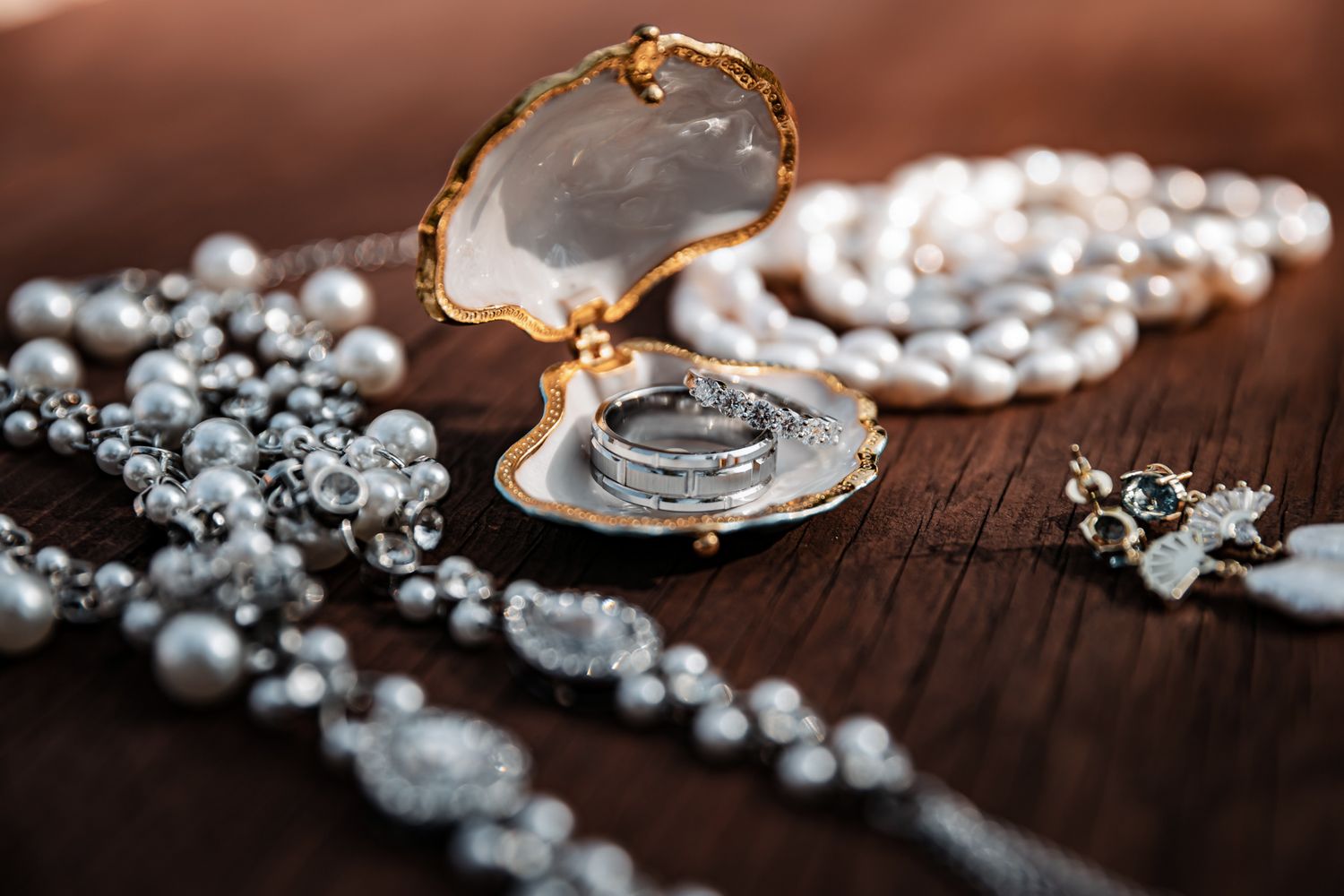 Silver wedding rings in an oyster shell jewelry box surrounded by pearl necklaces on a wooden surface.