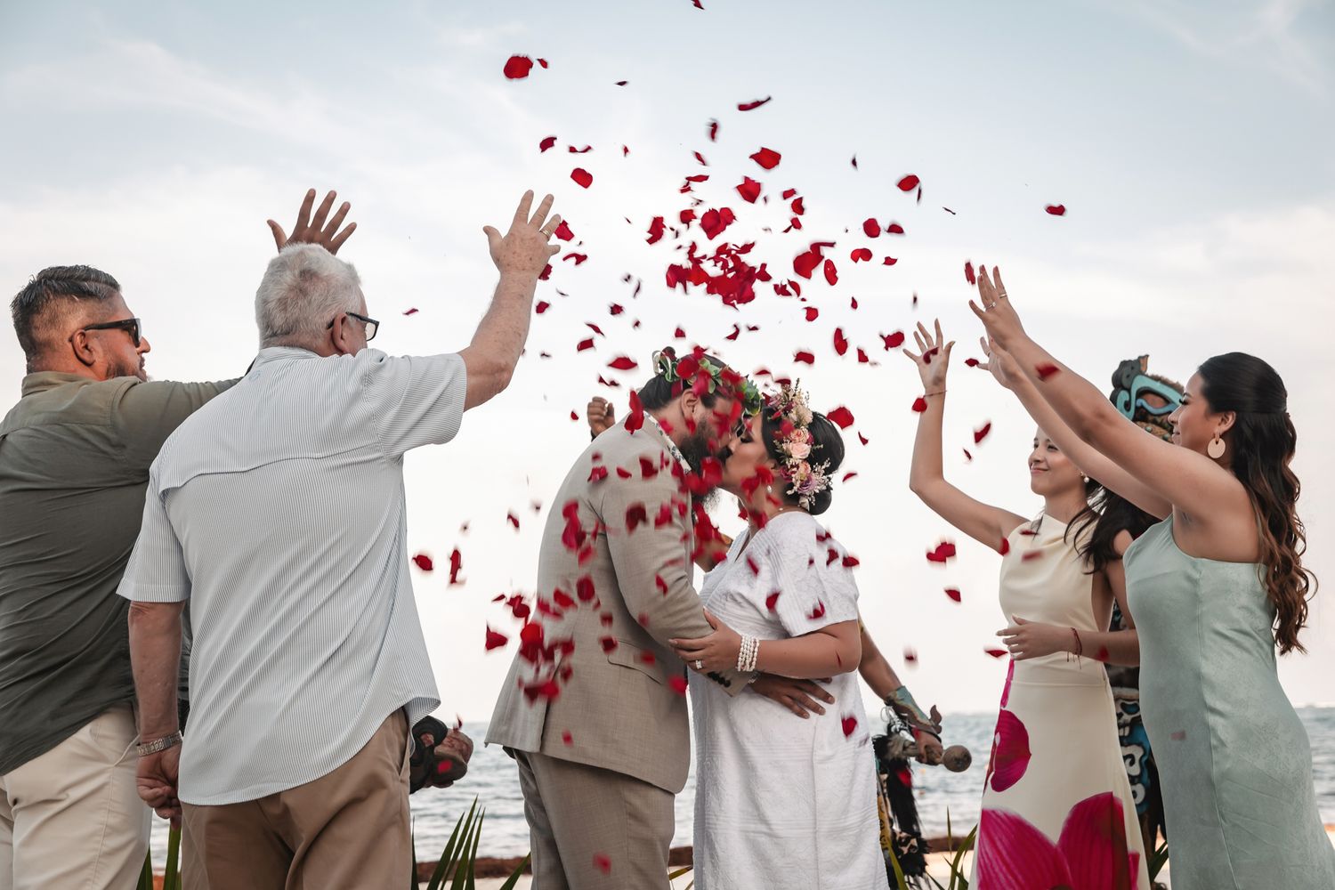 Couple kissing at beach wedding while guests toss red rose petals in celebration.