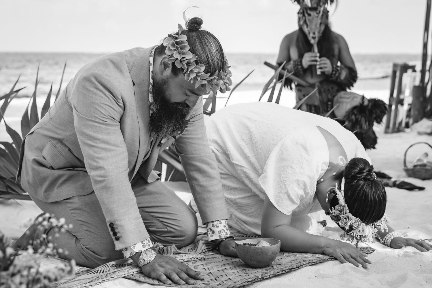 Couple kneeling on beach during traditional outdoor wedding ceremony with floral leis and cultural decorations.