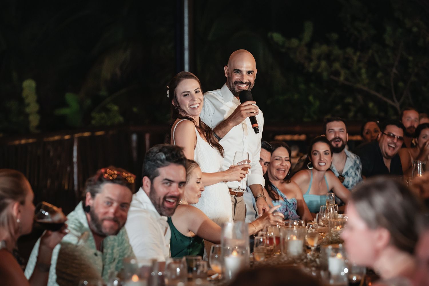 Couple shares a joyful moment with microphone at wedding reception dinner, surrounded by smiling guests at candlelit tables.