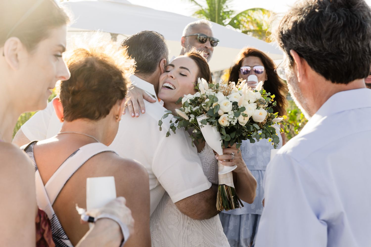 Bride in white dress hugging guests while holding white floral bouquet at outdoor wedding celebration.