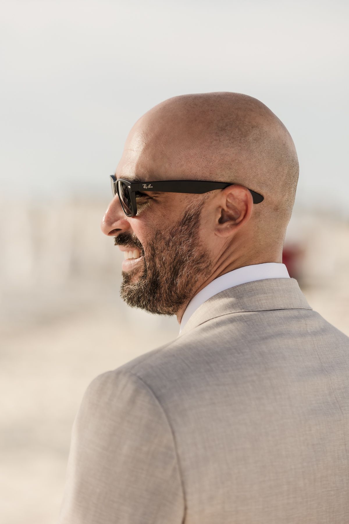 Bald man with beard wearing Ray-Ban sunglasses and beige suit laughing outdoors.