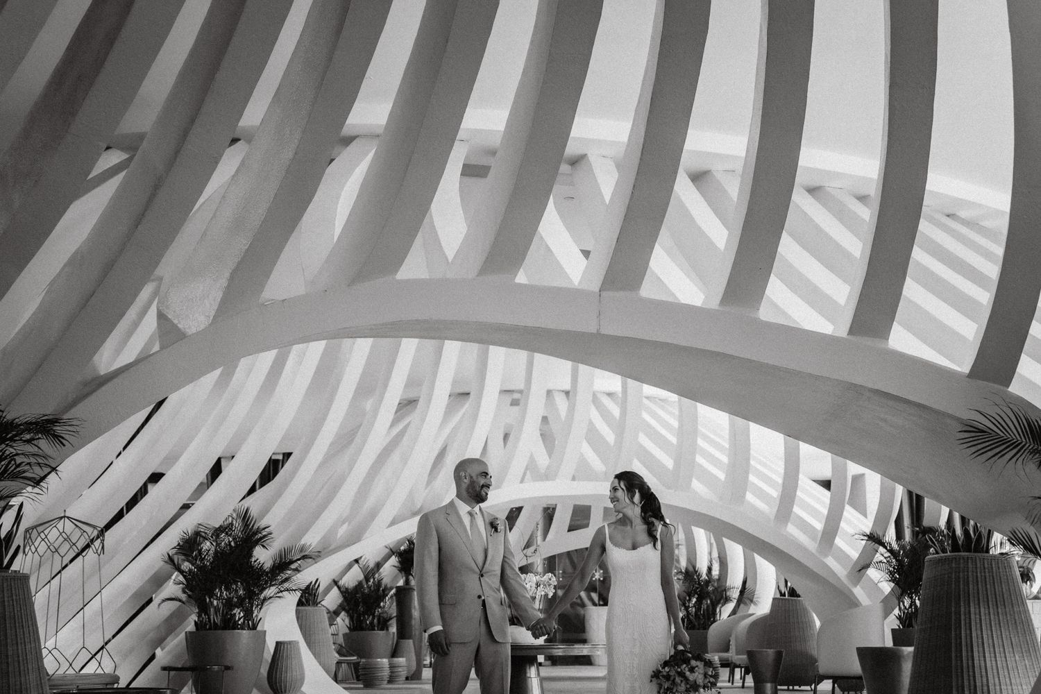 Black and white wedding photo of couple standing under elegant arched structure with sweeping curved lines at modern venue.