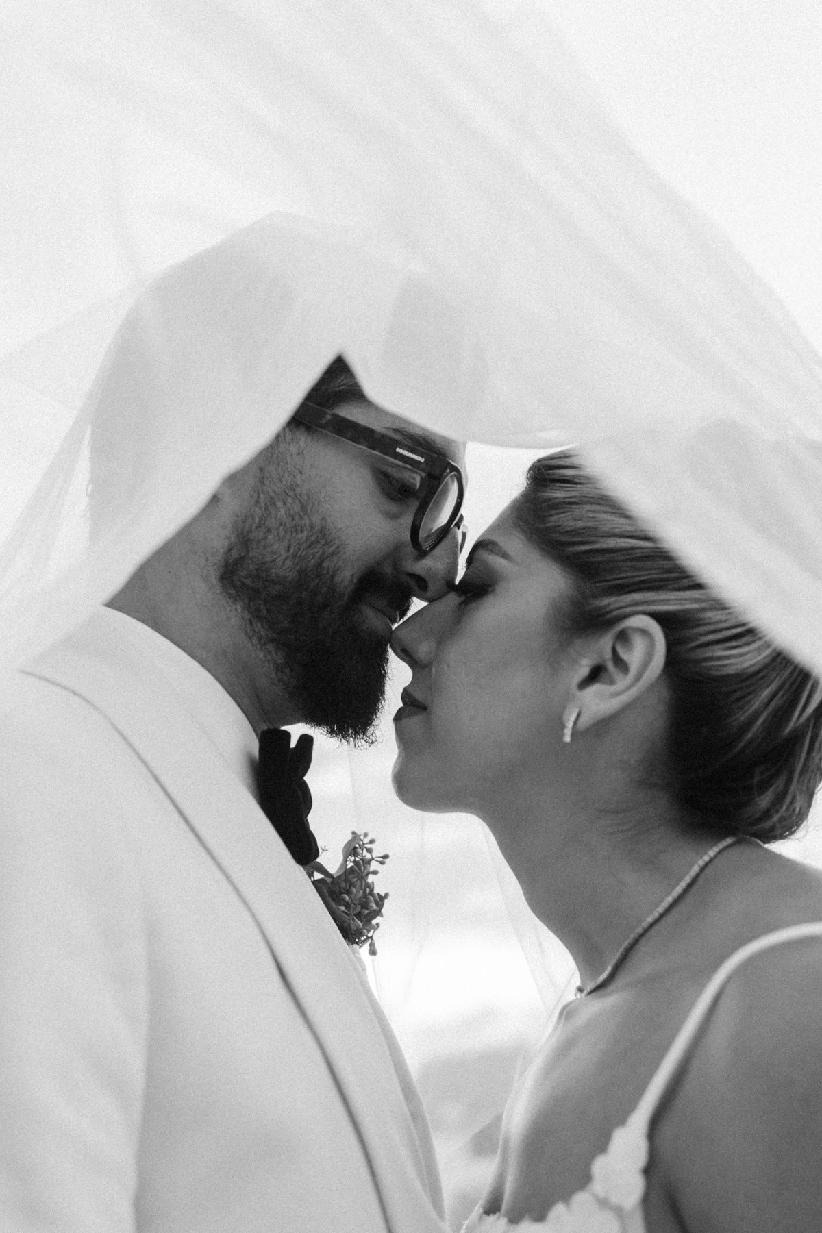 Black and white photo of bride and groom sharing a kiss under a flowing wedding veil.