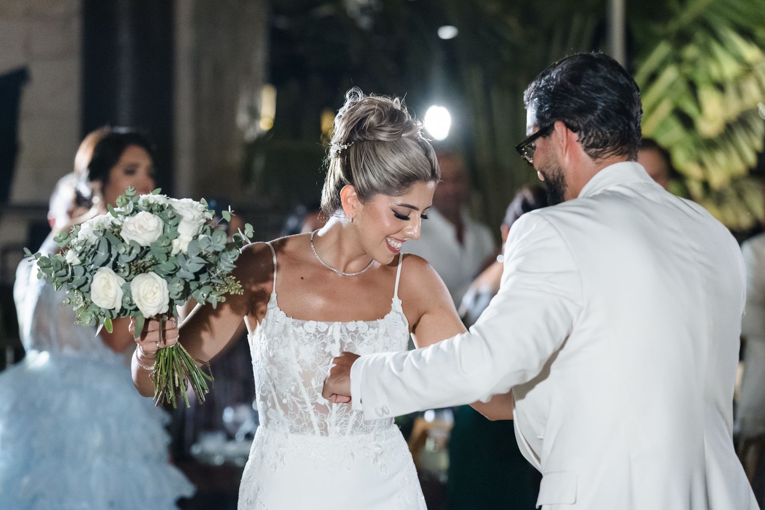 Bride in lace wedding dress holds white bouquet while groom in white suit adjusts her dress at outdoor wedding.