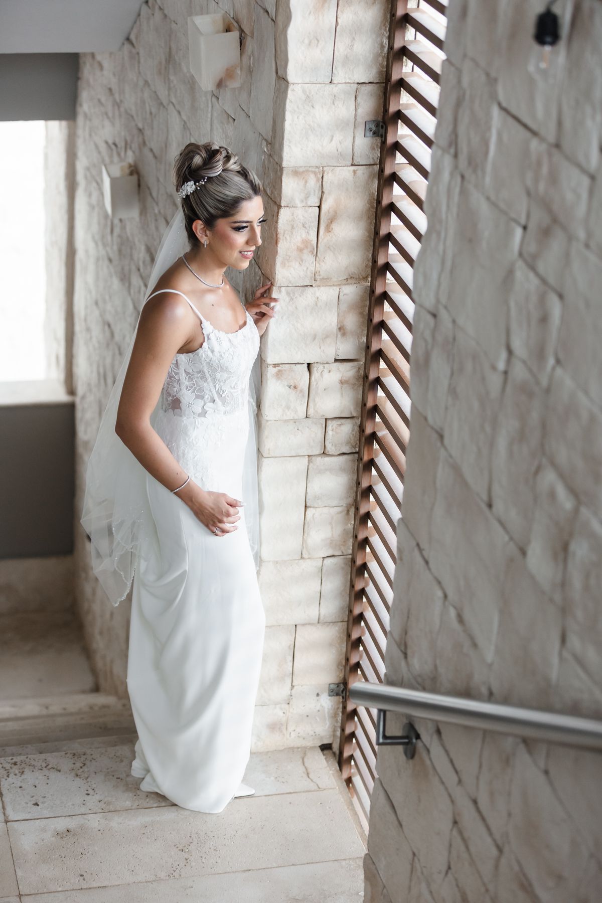 Elegant bride in white lace wedding dress standing by stone wall indoors.