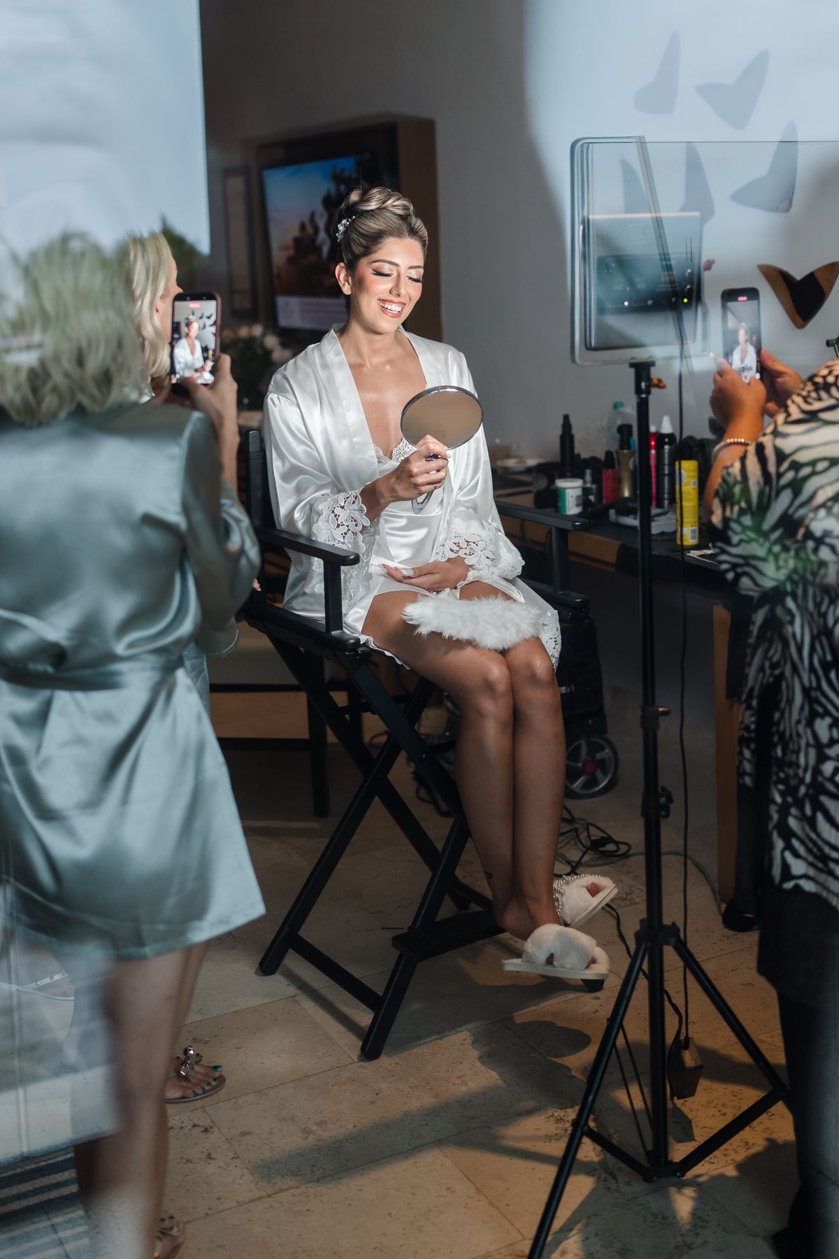 Bride in white satin robe sits in director chair holding mirror, smiling, while bridesmaids photograph her during wedding preparations.