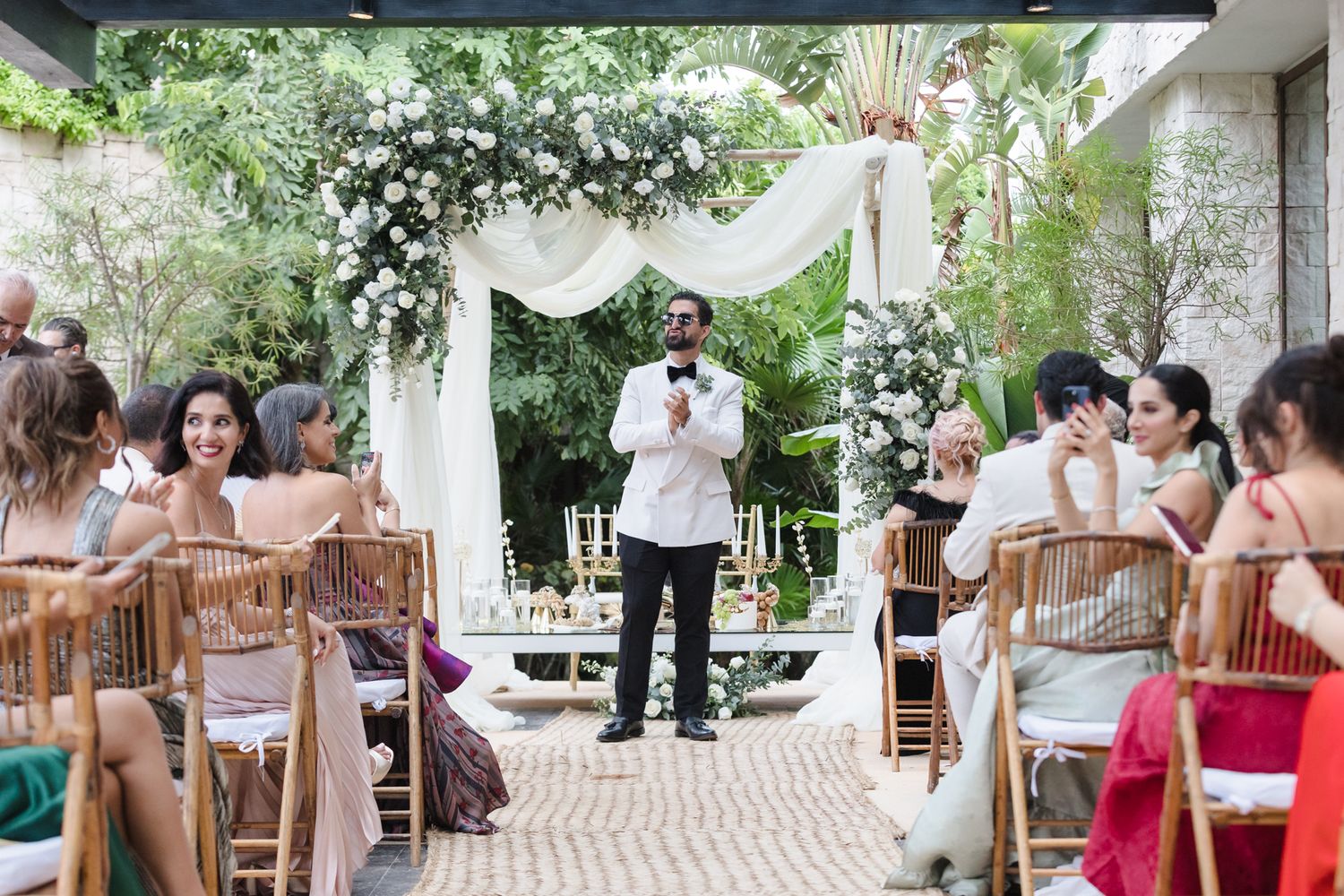 Man speaking at an elegant outdoor wedding ceremony with floral arch, seated guests, and lush greenery backdrop.