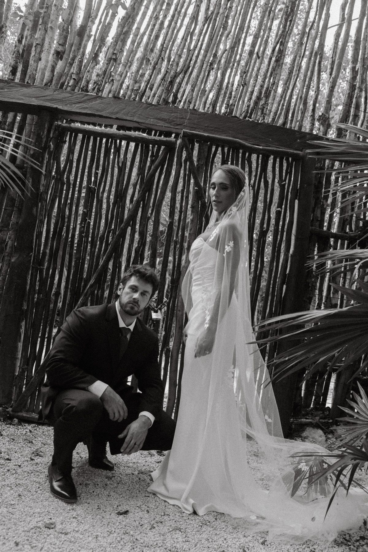 Black and white wedding photo of bride in veil gown and groom in suit posing by rustic bamboo structure outdoors.