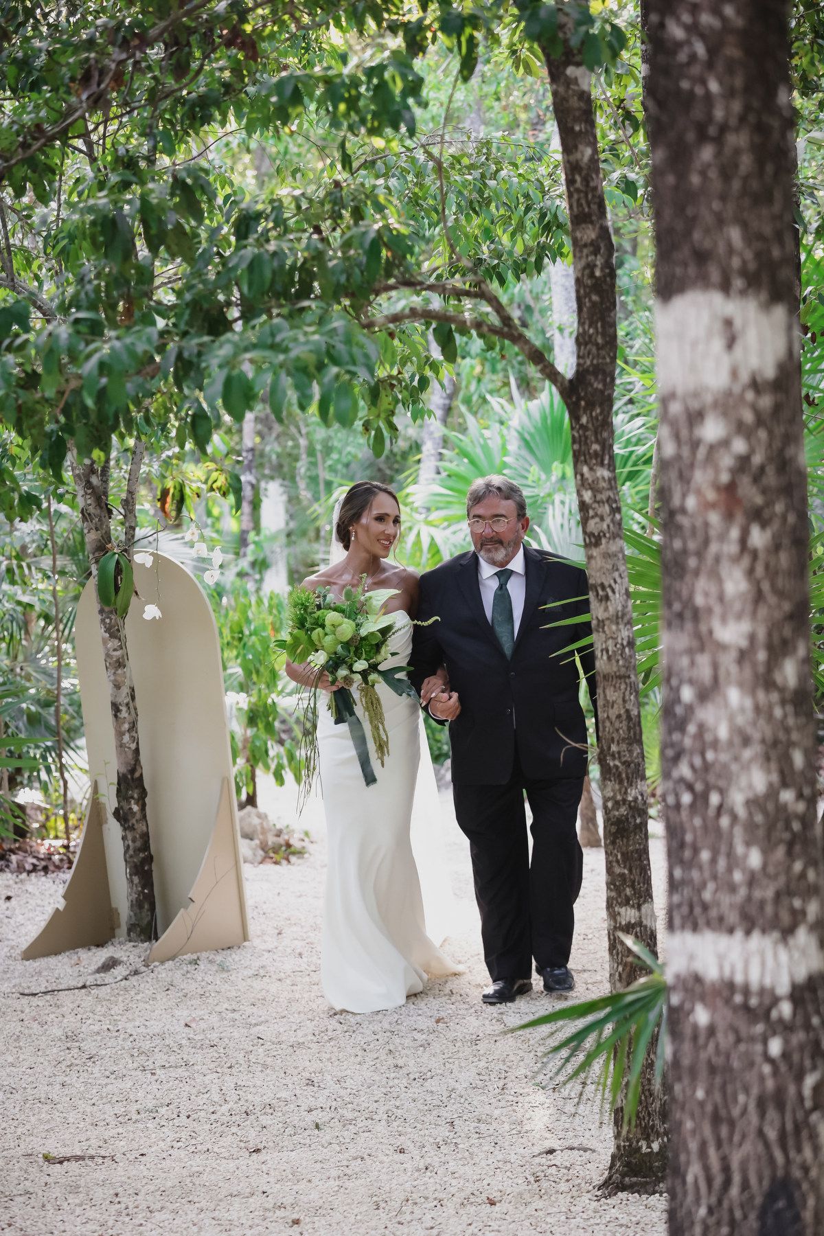 Bride in white gown walks with suited man through lush tropical garden path holding green bouquet.