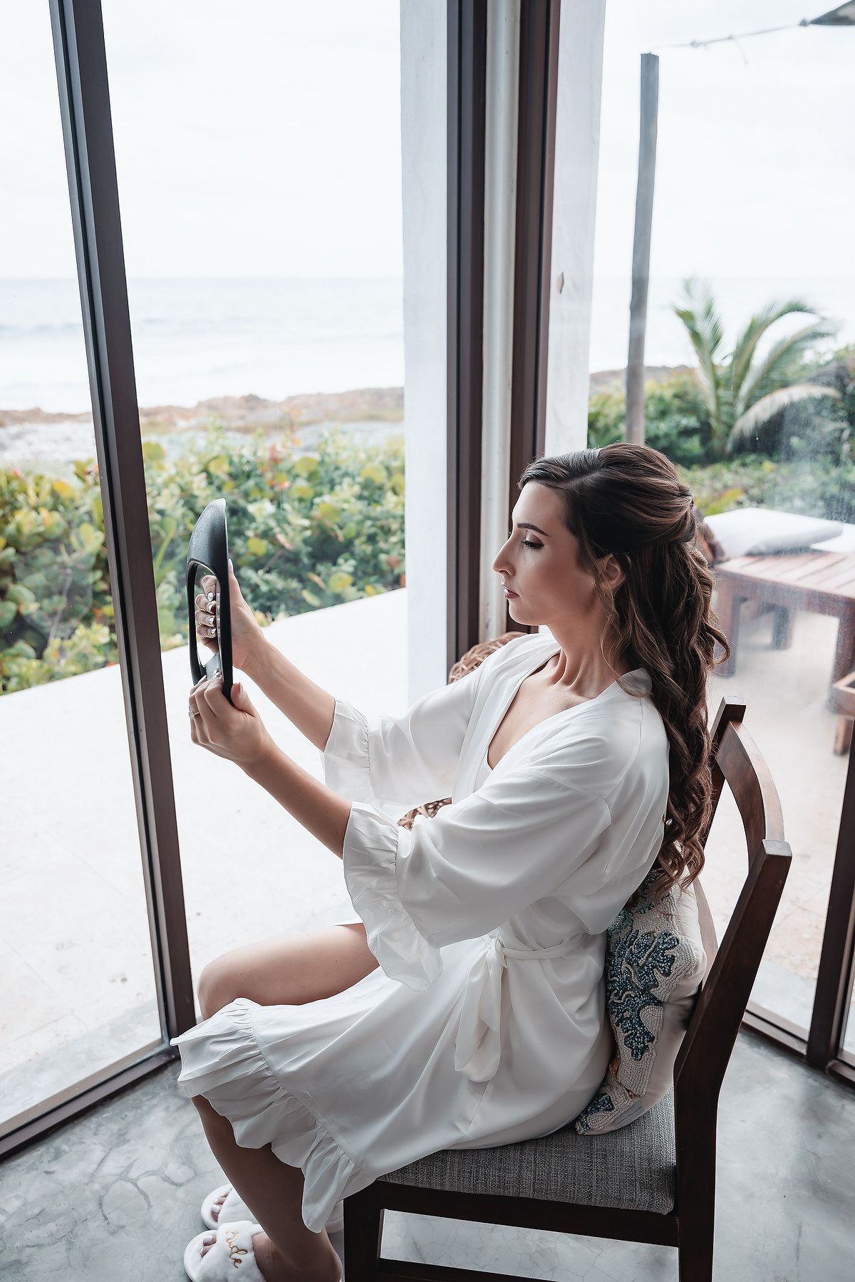 Bride in white robe sitting by window taking selfie with tablet, ocean view and palm trees in background.