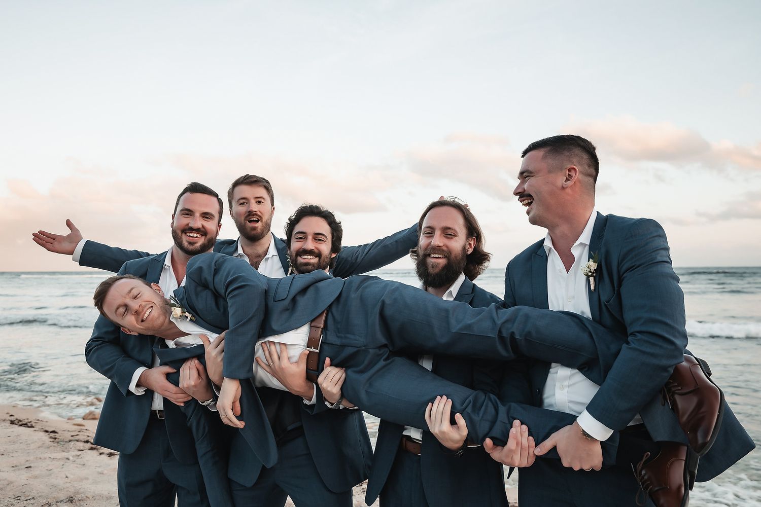 Groomsmen in navy suits carrying the groom on a beach at sunset, laughing and celebrating together.