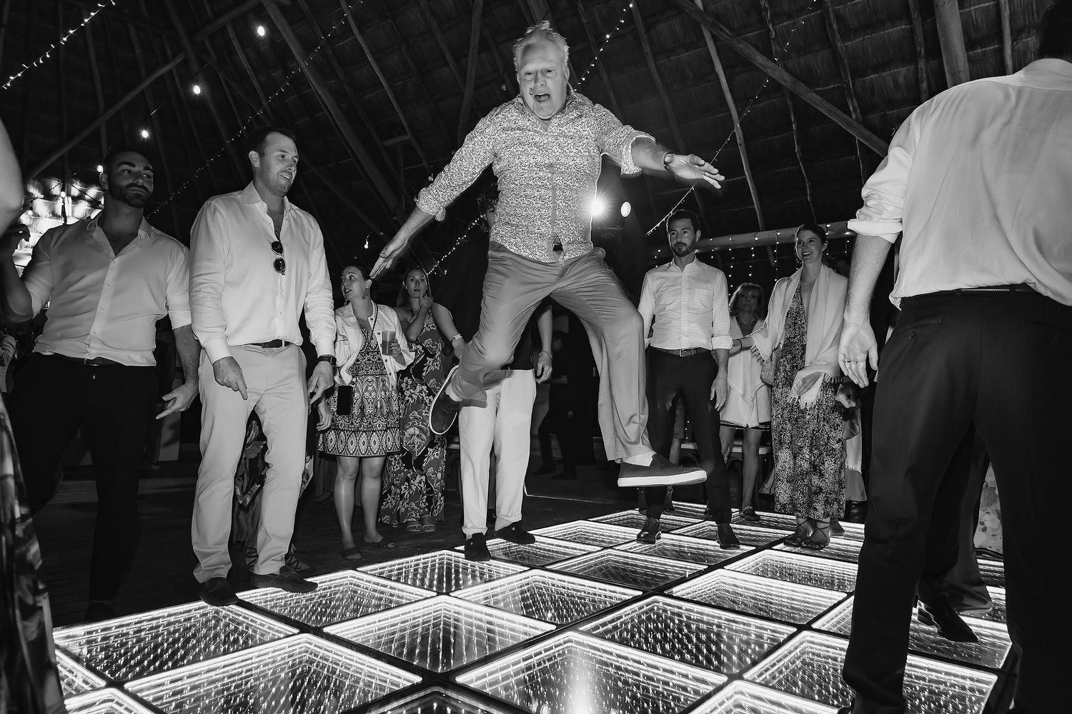 Man jumping on illuminated dance floor at wedding reception, surrounded by guests celebrating in black and white photo.
