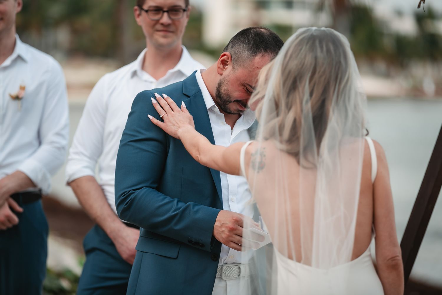 Emotional groom in blue suit crying as bride comforts him during heartfelt beach wedding ceremony vows.