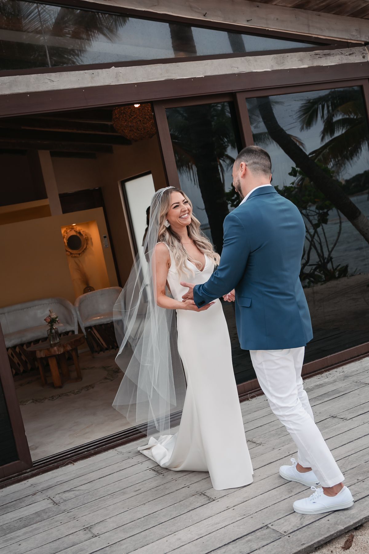 Bride in white gown and veil holds hands with groom in blue blazer on wooden deck at tropical wedding.