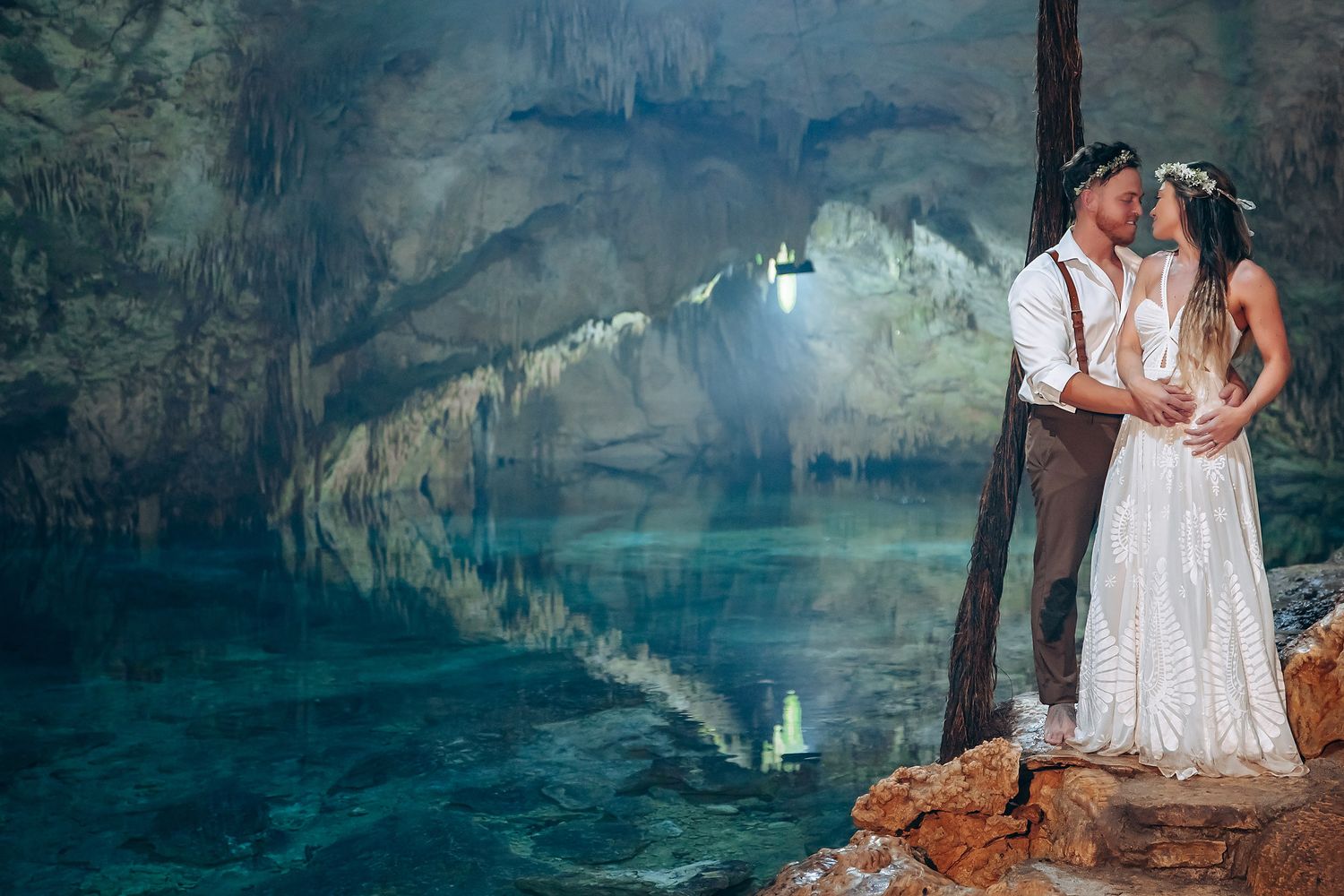Two figures in white attire standing near the edge of a stunning blue-green cenote in Mexico.