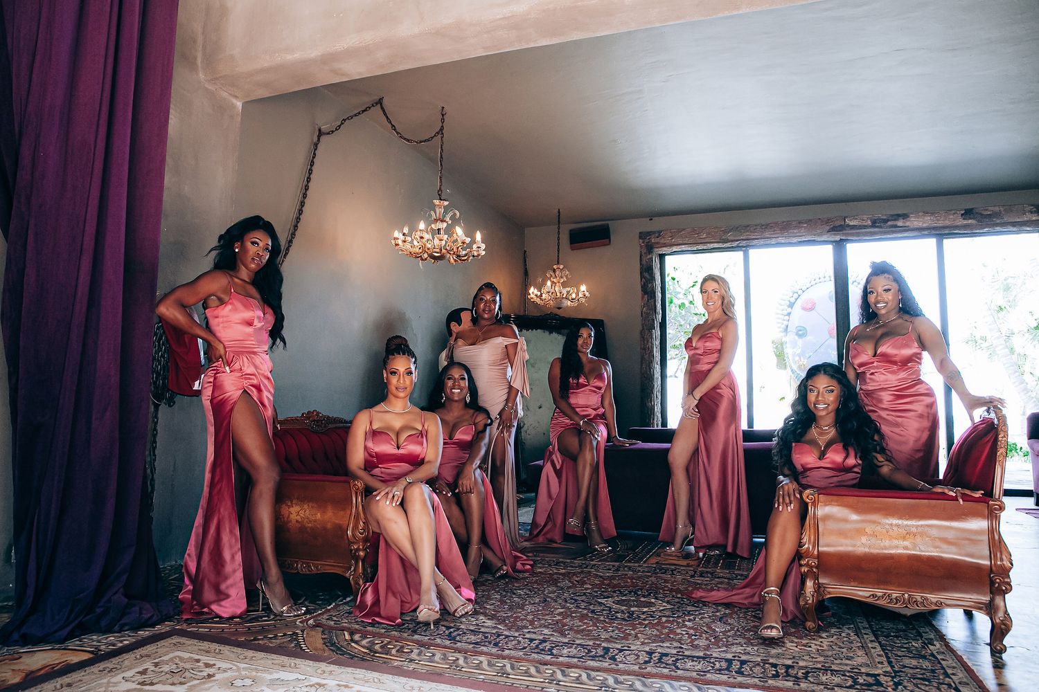 A group in matching pink satin dresses pose together in an elegantly decorated room with a chandelier and curtains.