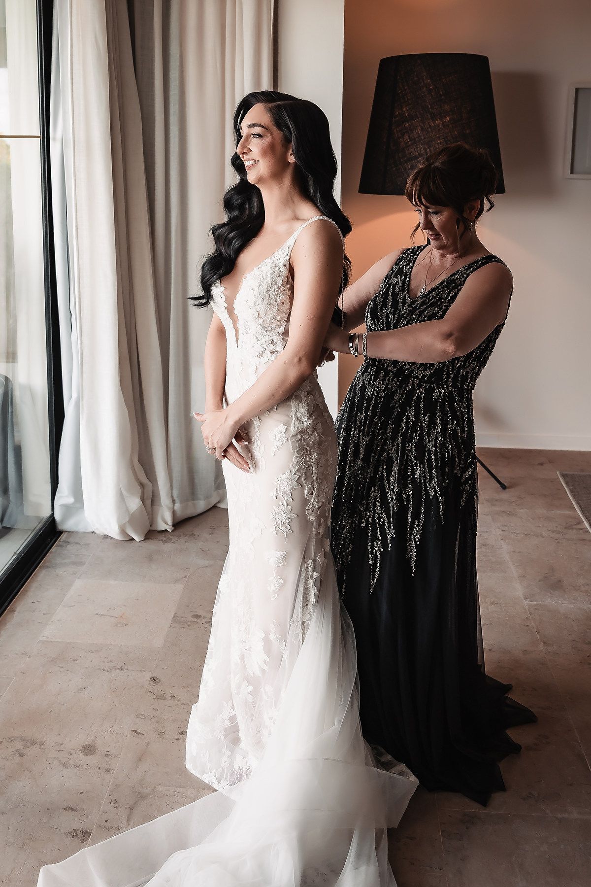 A bride in a white lace gown stands with attendant in black dress during pre-wedding moments.