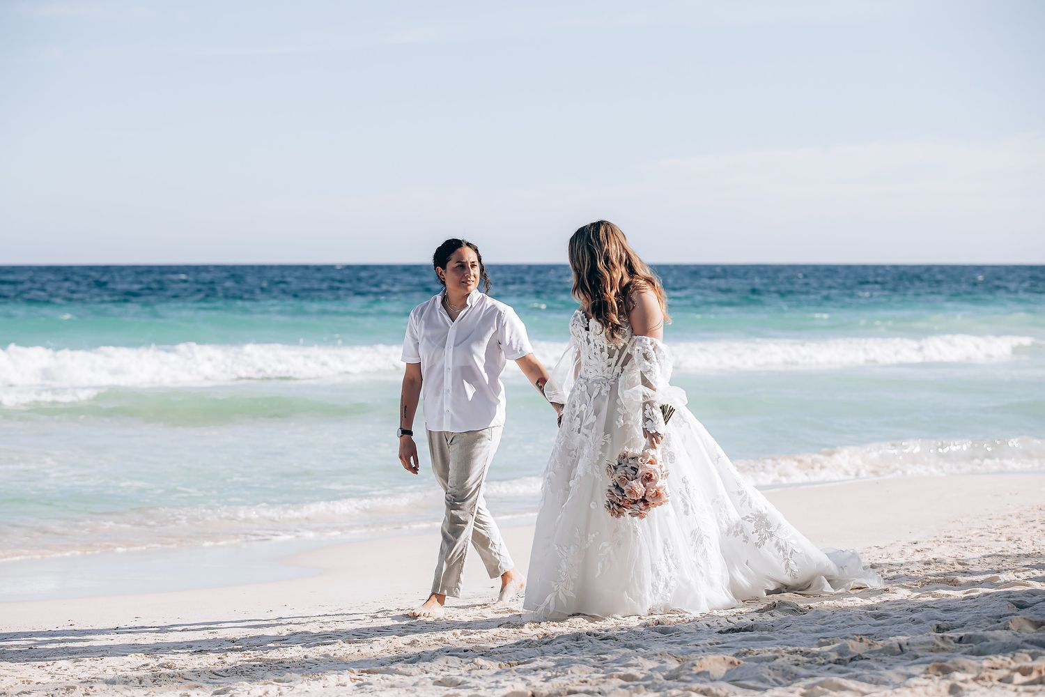 A couple in white attire runs playfully along a pristine beach with turquoise waters and white sand.