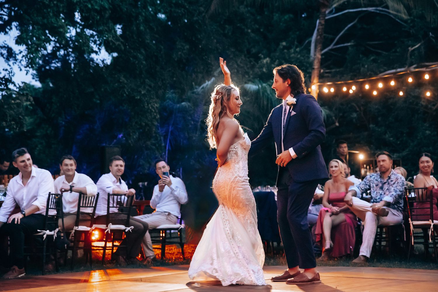 Evening wedding reception under string lights with guests seated at outdoor tables.